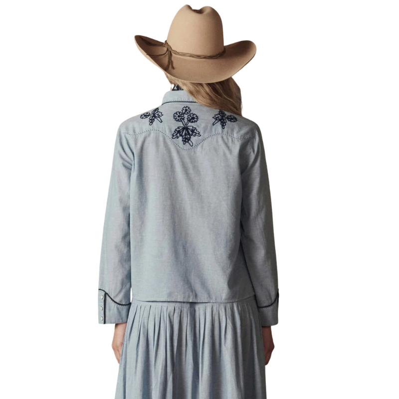 Person wearing a light blue denim jacket with floral embroidery and a beige cowboy hat on a white background