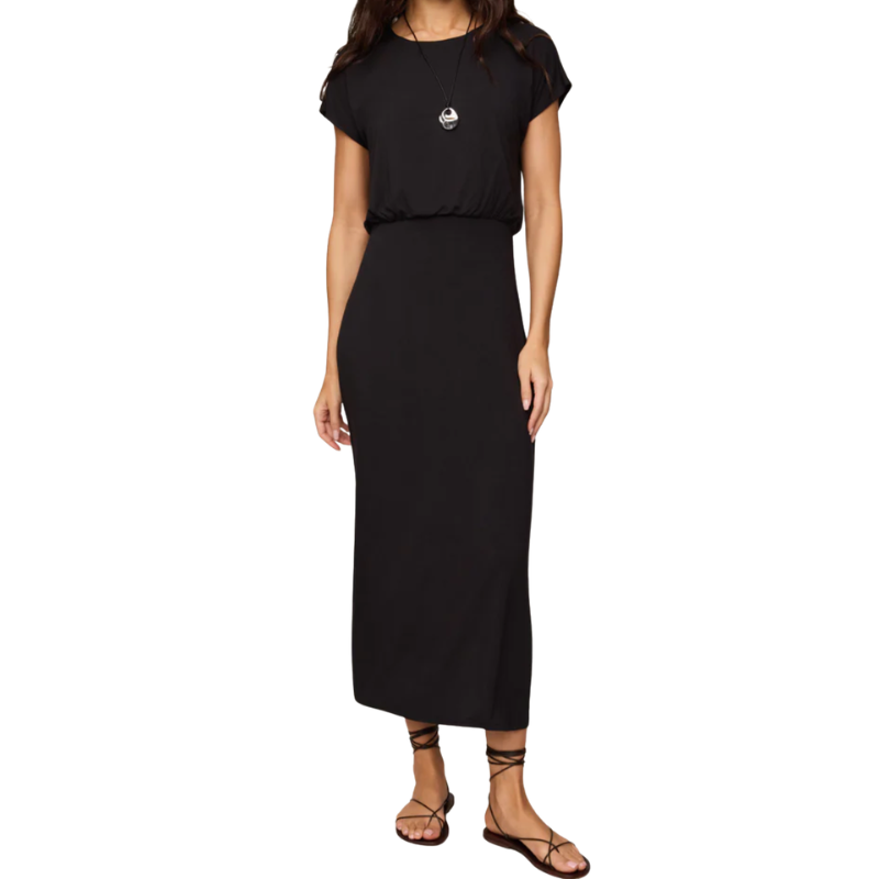 A woman modeling a black short-sleeve midi dress with a small white graphic on the chest, paired with black strappy flat sandals, against a plain white background.