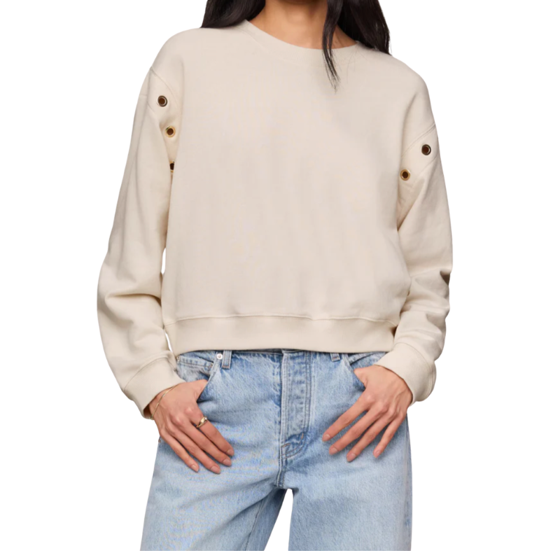 A woman wearing a cream-colored crewneck sweatshirt with decorative brown buttons on the sleeves, paired with light blue jeans, shown against a plain white background.