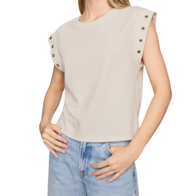 A woman wearing a light beige sleeveless top with decorative metallic snap buttons on the shoulders, paired with light blue jeans, against a plain white background.