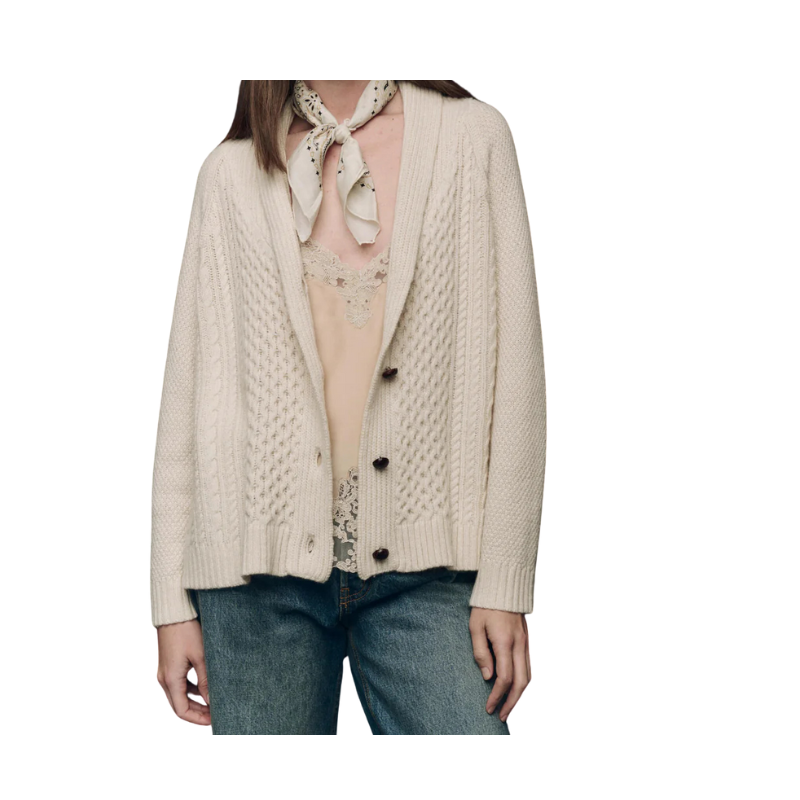 Person wearing a beige cable knit cardigan over a lace top and jeans on a white background