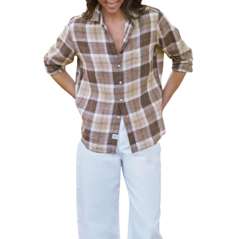 A woman wearing a brown and beige plaid button-down shirt with rolled-up sleeves and white pants, shown from the waist up against a plain white background.