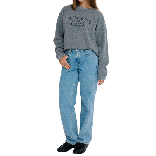 Person wearing a gray sweatshirt with 'Homebody Club' text and blue jeans on a white background