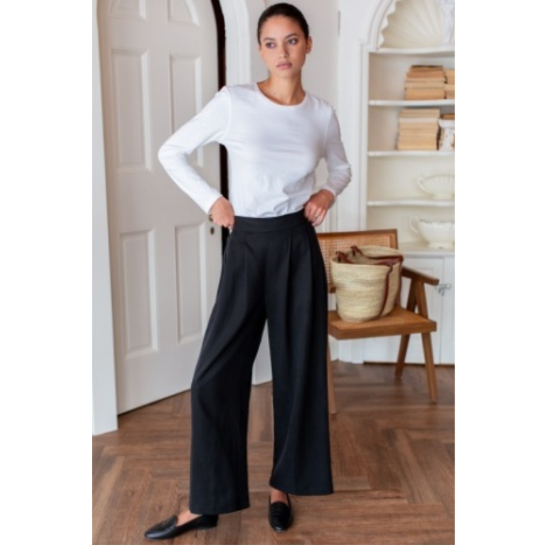 Woman wearing a white long-sleeve top and black wide-leg pants in a room with wooden floor and white walls.