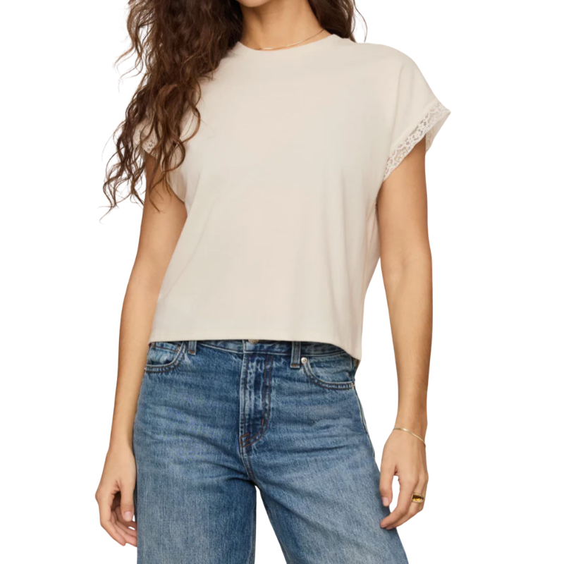 A woman wearing a light beige short-sleeve crop top with delicate lace trim on the sleeve cuffs, paired with blue jeans, against a plain white background.