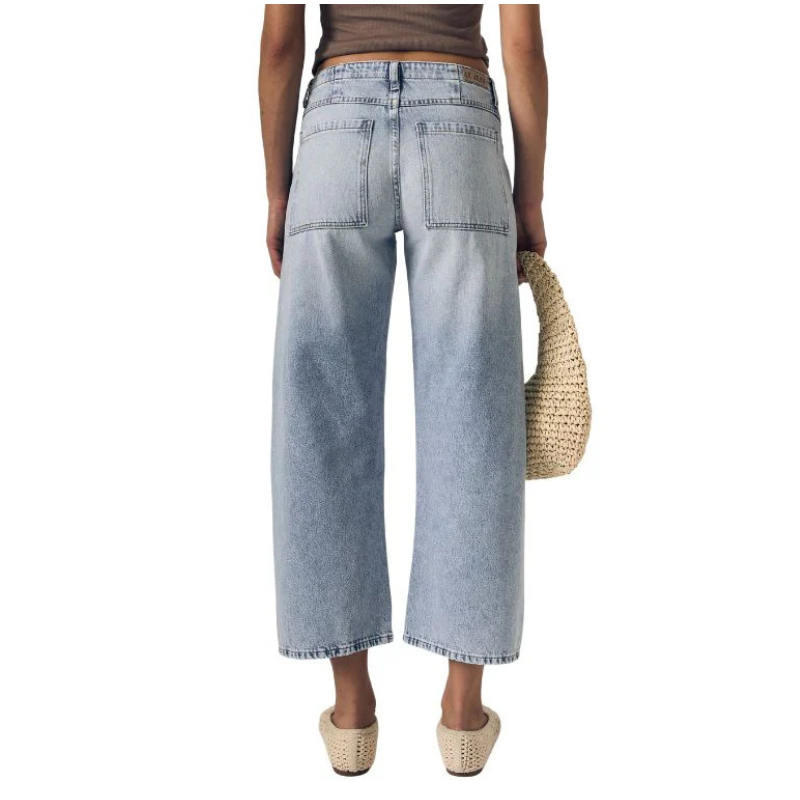 Person wearing light blue wide-leg jeans holding a straw bag on a white background