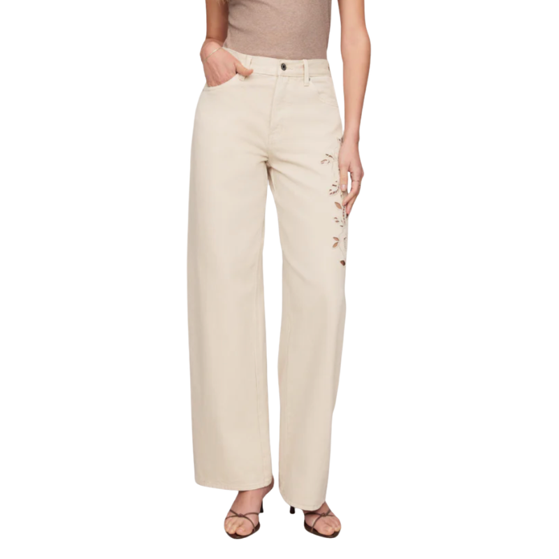 A model wearing a pair of wide-leg beige pants with floral embroidery on the side, paired with a beige top and brown strappy heels, shown against a plain white background.