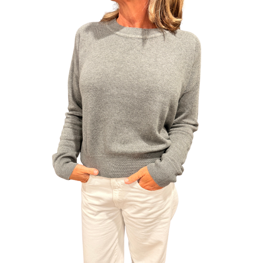 A woman wearing a light gray knit crewneck sweater and cream-colored pants, standing against a plain white background.